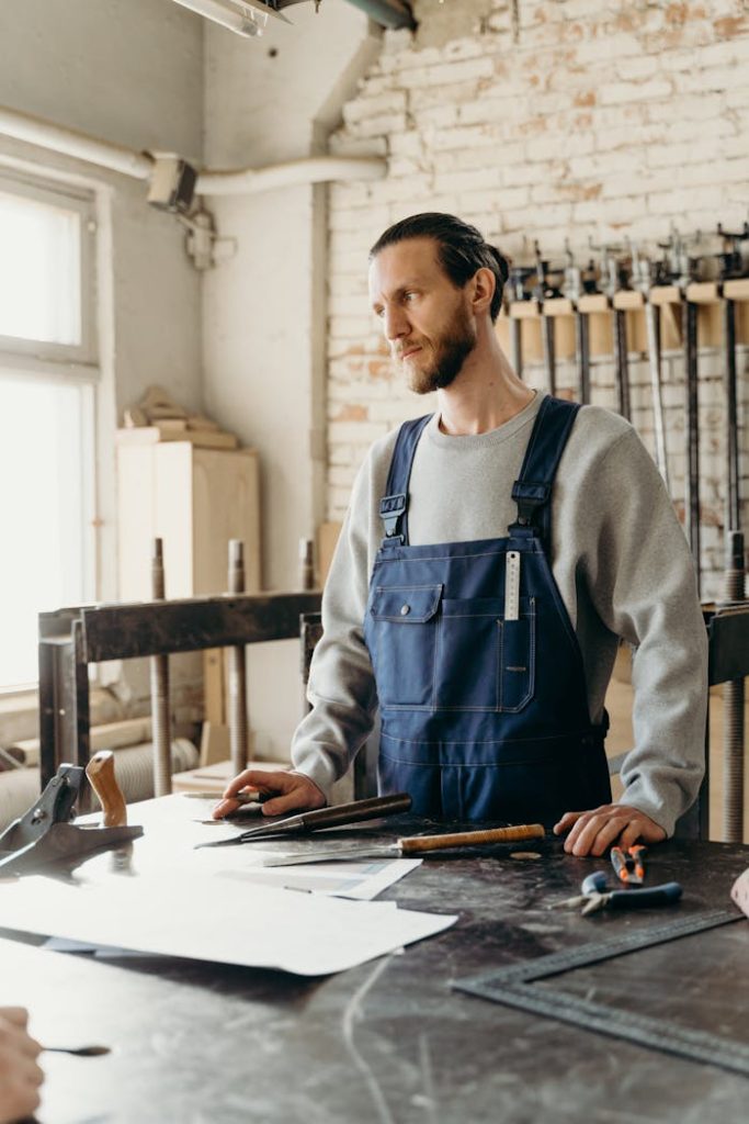 Caucasian craftsman in a workshop wearing overalls, surrounded by tools, looking focused.
