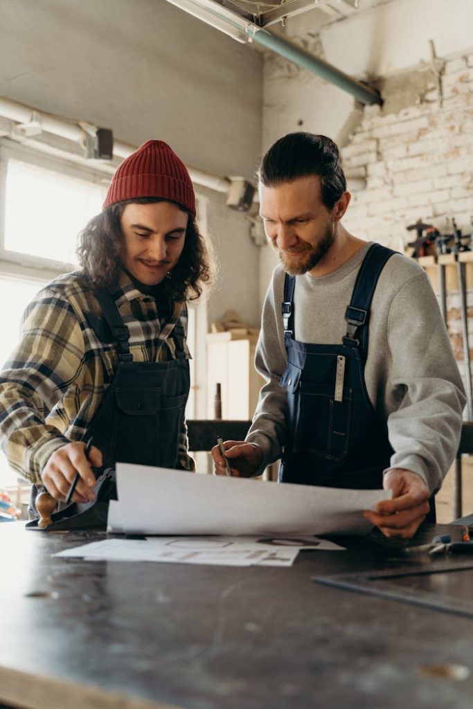 Two carpenters in overalls plan a project in an industrial workshop. Room is filled with tools and natural light.